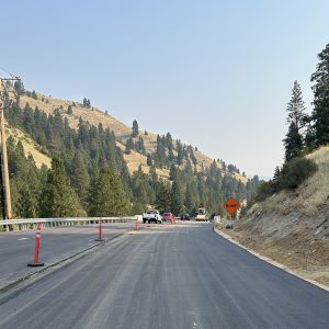Fresh pavement on SH-55 near the Fleming Creek Bridge, with traffic cones, construction vehicles and a “Bump” warning sign marking the active work zone in a mountainous, forested area.