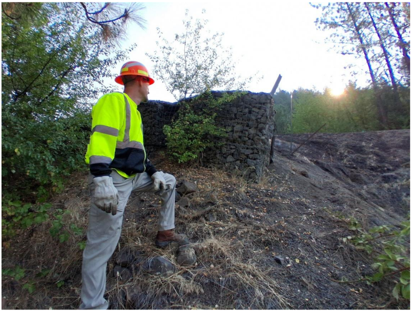 An ITD employee in high-visibility safety gear stands on a steep hillside near SH-11, inspecting the slope and an existing rock retaining structure along the Greer Grade.