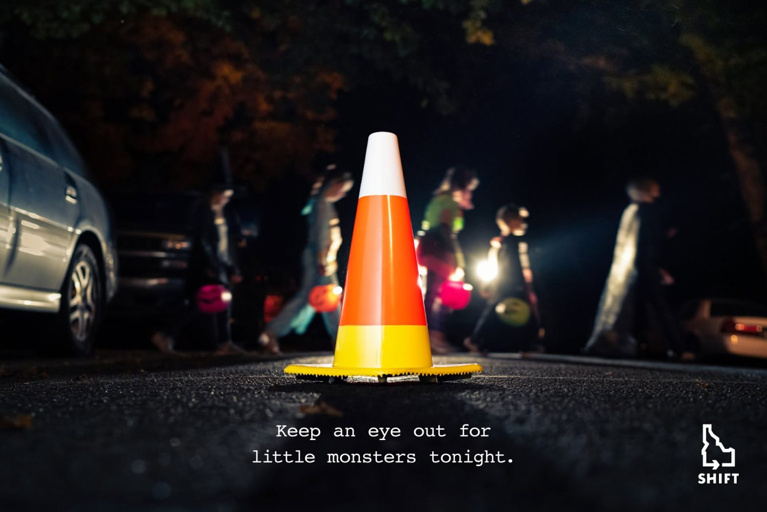 A traffic cone painted to look like candy corn on a dark street with children in costumes in the background. Text says "Keep and eye out for little monsters tonight."